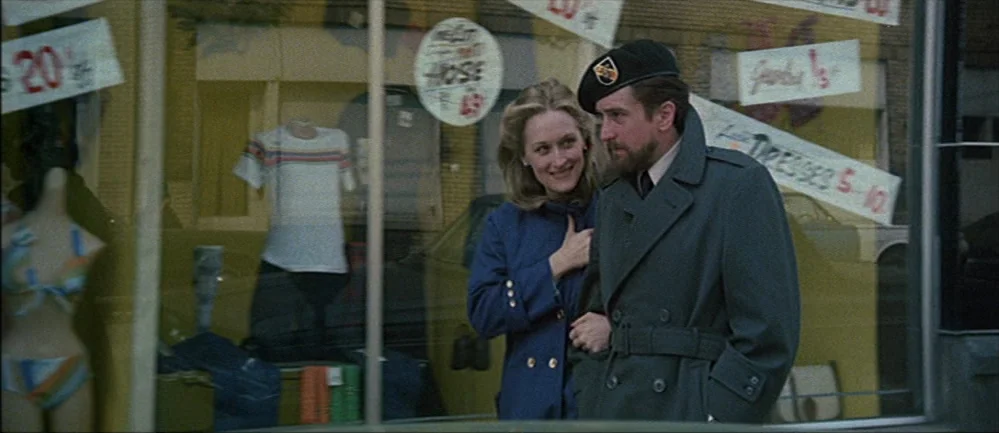 The Deer Hunter (1978) movie still: medium — A man in a military coat and a woman in a blue jacket walk together. They are passing a storefront…
