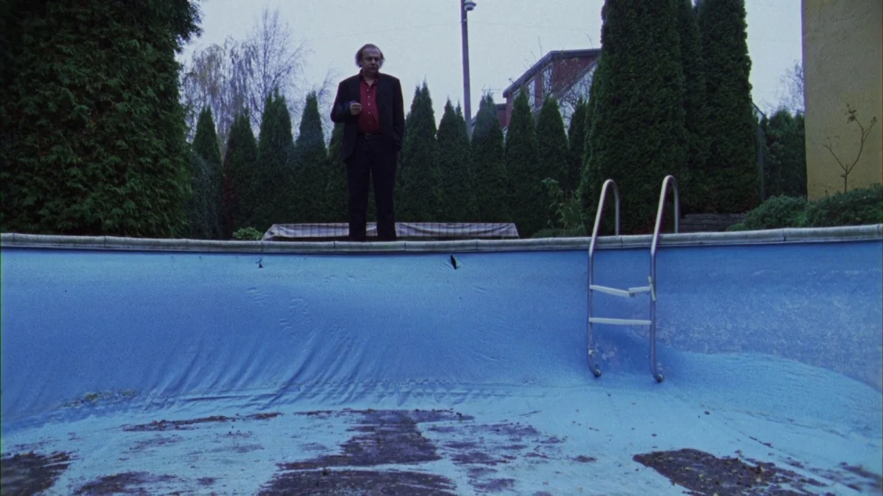 Pusher III (2004) movie still: wide — A suburban backyard features an empty blue swimming pool surrounded by tall evergreen hedges. A man in a dark…