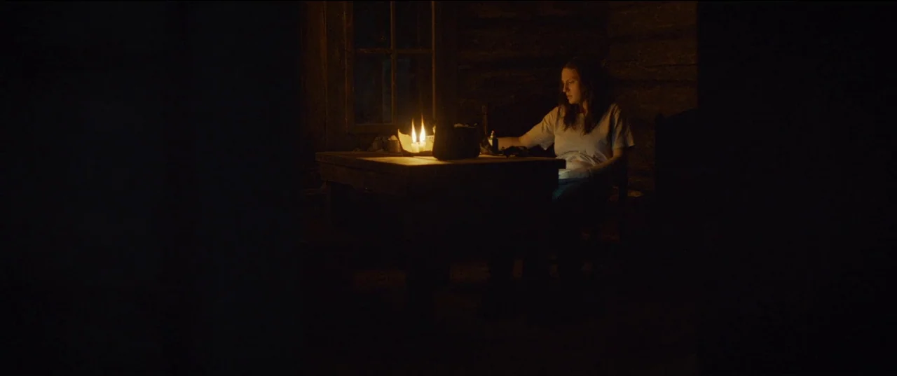Nightsiren (2022) movie still: medium — A dark, wood-paneled room interior features a woman sitting at a central table. She observes objects placed…