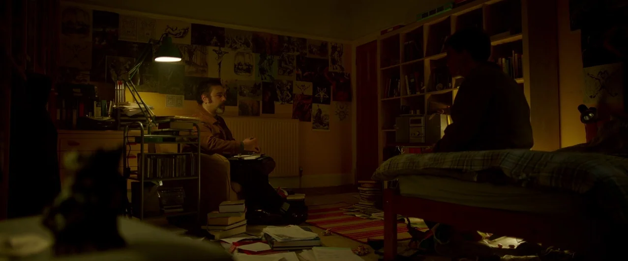 Ghost Stories (2017) movie still: wide — A dimly lit room filled with books, papers, and numerous occult posters on the walls. An adult male sits in…