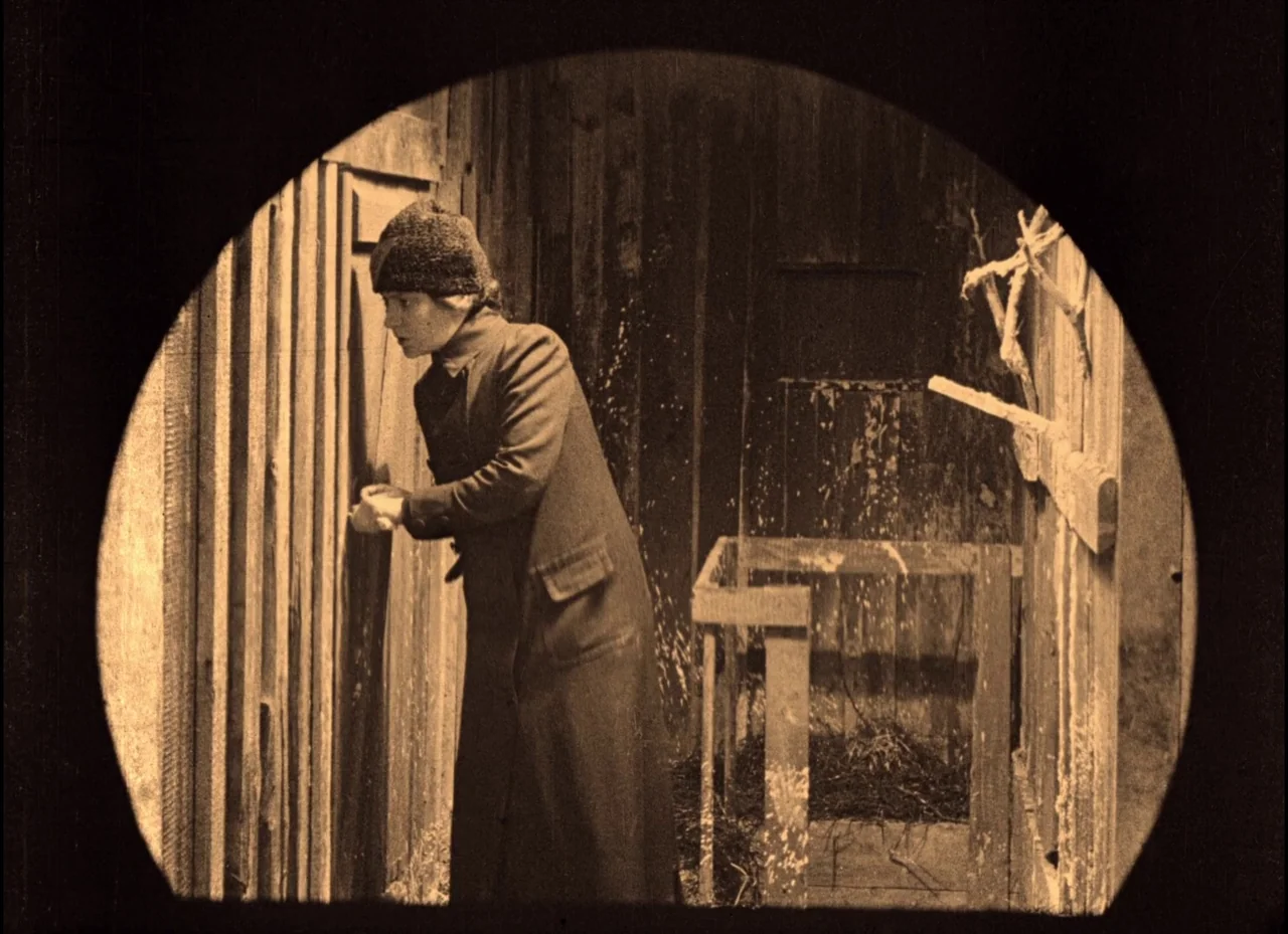 The Phantom Carriage (1921) movie still: medium — A dimly lit wooden structure with vertical planks and straw-covered floor. A woman in a dark coat…