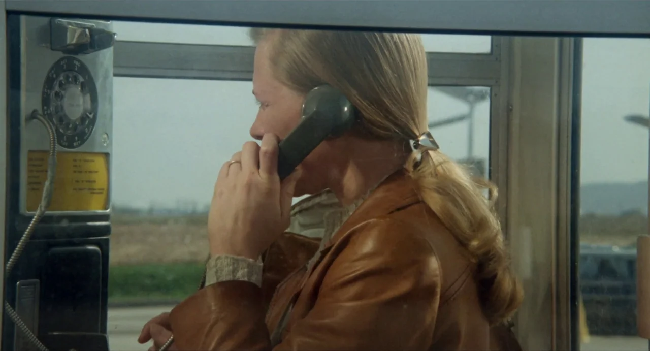 The Rain People (1969) movie still: medium — A glass-walled booth containing a rotary phone and a blonde woman. The woman stands inside while…