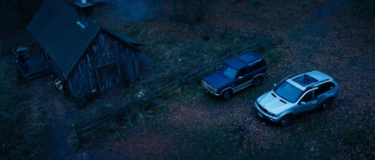 The Descent (2005) movie still: establishing — A dark, wooden cabin sits in a clearing surrounded by woods covered in fallen leaves. Two SUVs are…