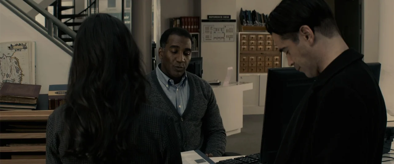 Winter's Tale (2014) movie still: medium — A quiet library reference desk contains documents, computers, and wooden cabinets with index drawers.…