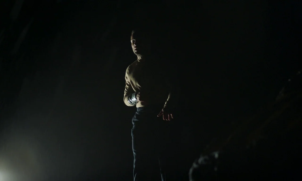 It Is In Us All (2022) movie still: medium — A dark, undefined exterior space contains only a single figure in the frame. The man moves through the…