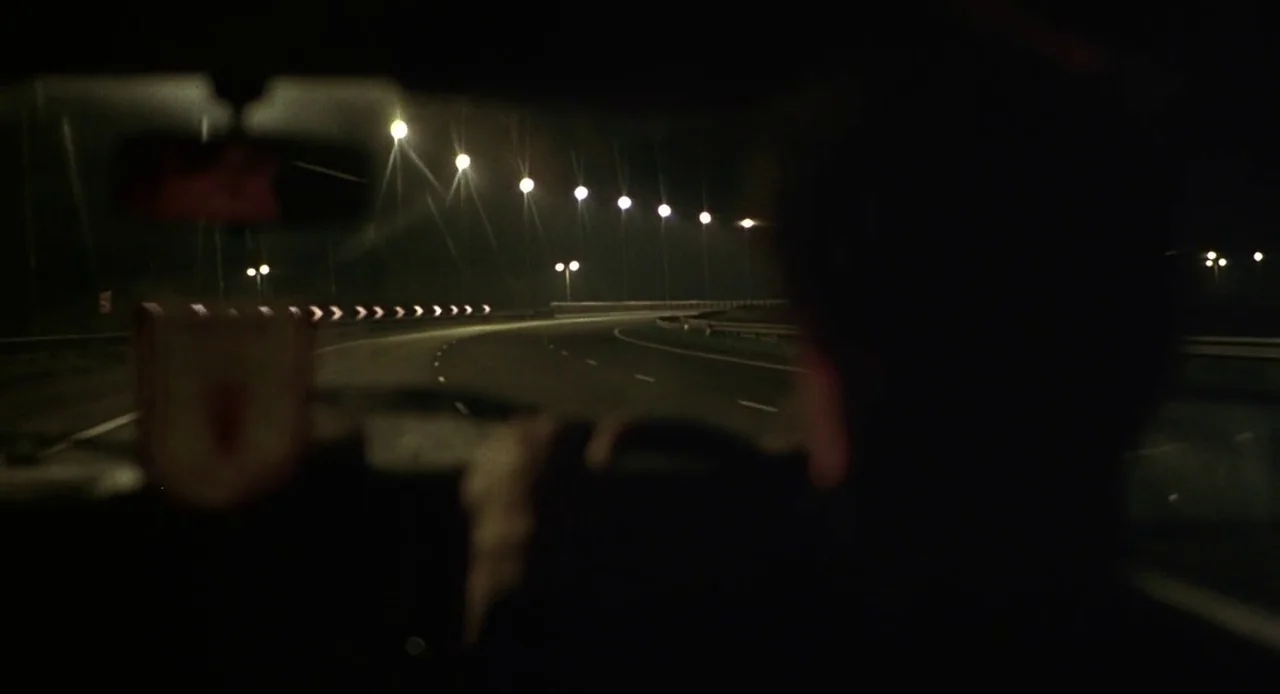 Naked (1993) movie still: medium — A view from inside a car moving along a dark, multi-lane highway at night. Rows of illuminated streetlights…