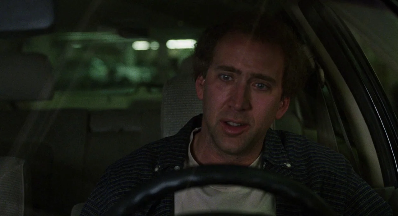 Adaptation (2002) movie still: medium — A man sits in the driver seat of a vehicle at night. The background shows blurred lights of a parking…