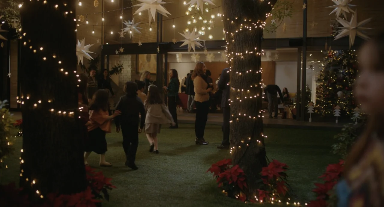 Dating Santa (2023) movie still: wide — A festive outdoor patio gathering features guests illuminated by hanging string lights and stars. Children…