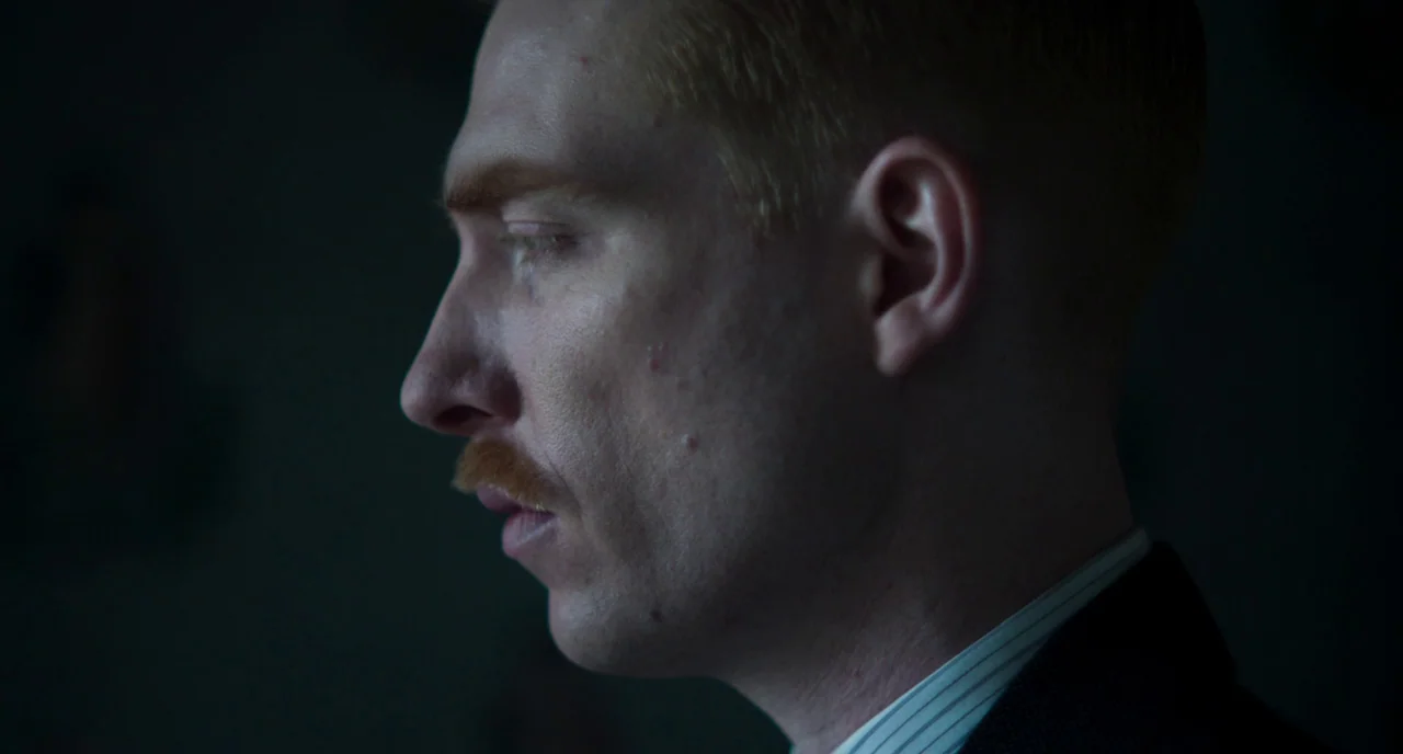 The Little Stranger (2018) movie still: closeup — A man in a suit is seen in extreme profile against a dark background. He stands facing left with…