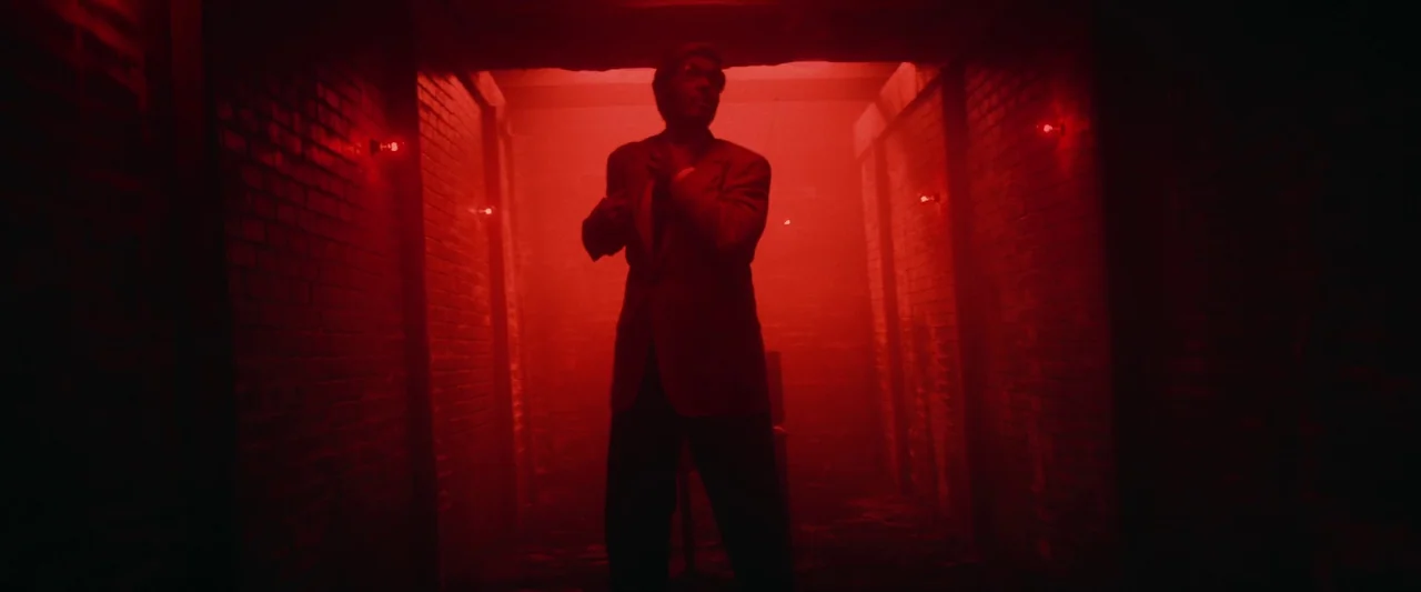 Revealer (2022) movie still: fullbody — A narrow brick hallway is saturated in deep red light from small wall fixtures. A man stands in the center…