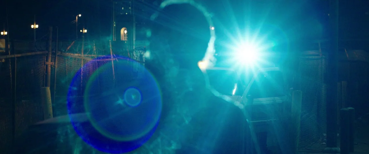 Nightcrawler (2014) movie still: medium — An outdoor parking area at night is enclosed by a chain-link fence with barbed wire. A person stands…