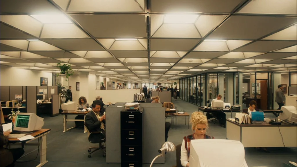 The Informant! (2009) movie still: wide — A large open-plan office features rows of workstations with beige cubicle walls and computers. People are…