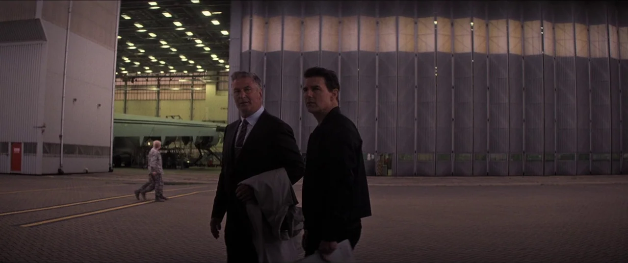 A massive industrial aircraft hangar features sliding accordion metal doors and concrete flooring. Two men walk across the tarmac in the foreground while a third figure walks behind.