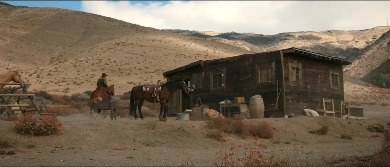 Dead for a Dollar (2022) movie still: wide — A dry, hilly landscape featuring a rustic dark wood structure with multiple windows. A person on…