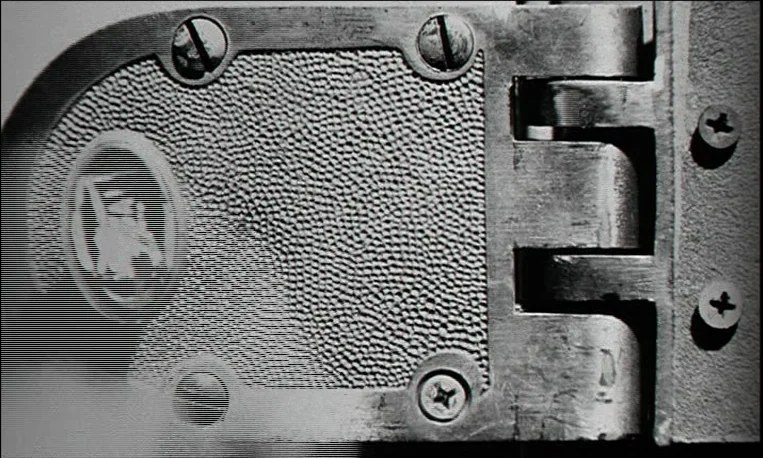 Pi (1998) movie still: closeup — A close-up view of a metal door hinge with a textured decorative plate. The hinge mechanism is secured to the frame…