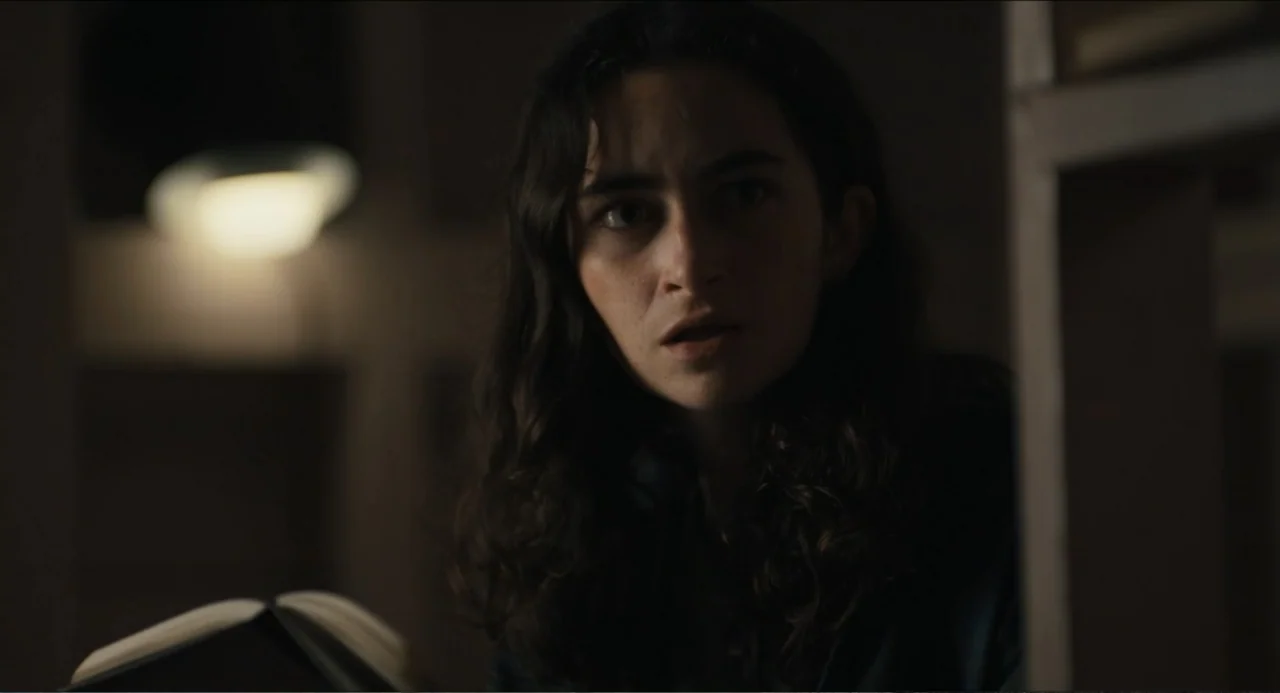 Hell of a Summer (2025) movie still: closeup — A dimly lit interior space features bookshelves partially obscured in the deep shadows. A young woman…