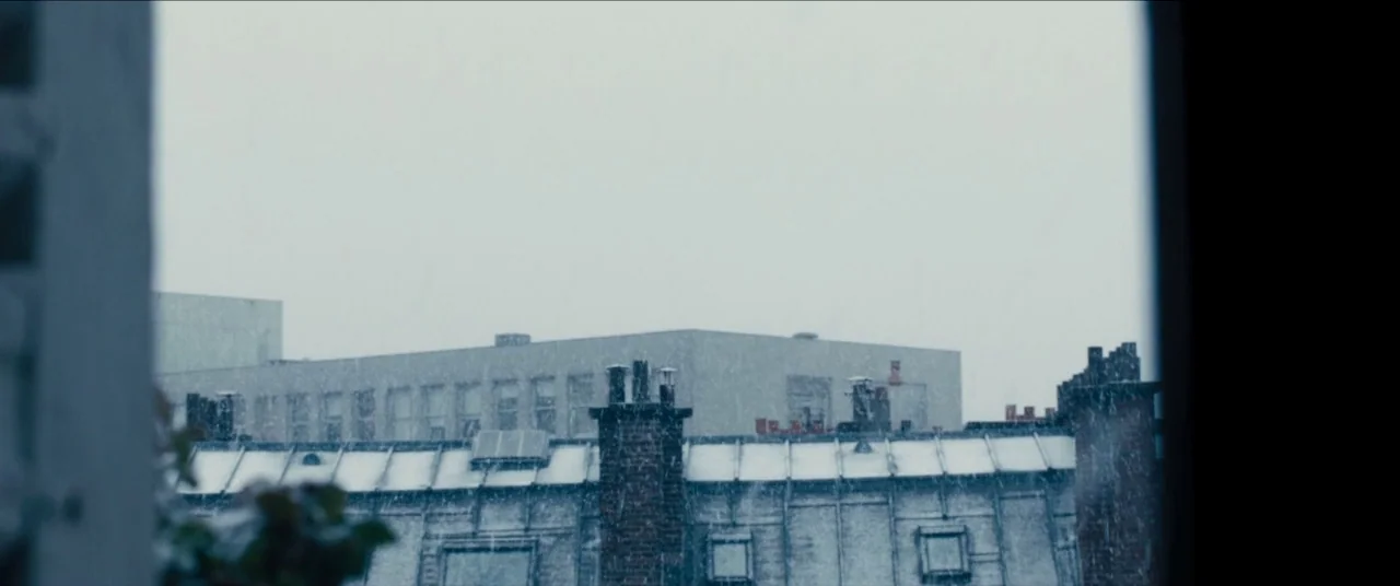Simon Killer movie still: establishing — An urban rooftop landscape is blanketed in light snow during a grey, overcast day. Brick chimneys and…
