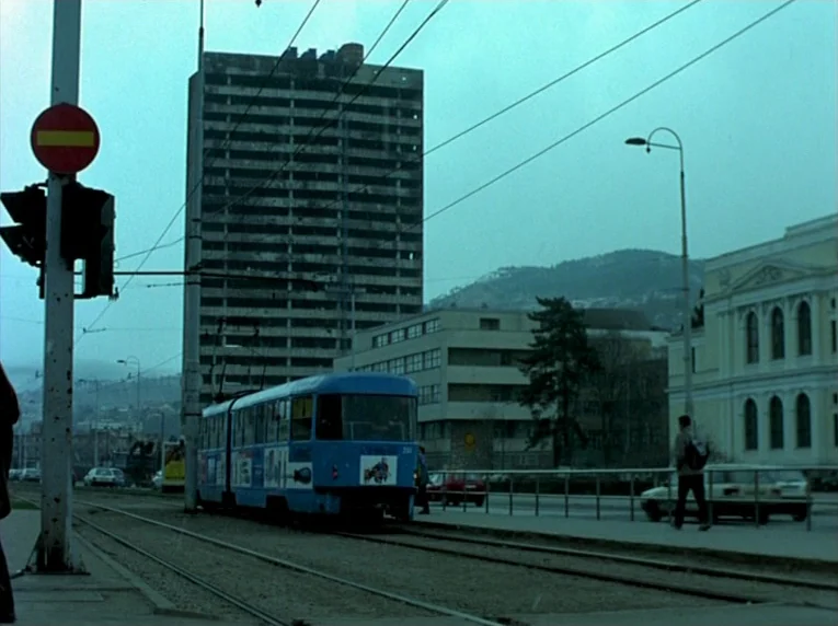 Notre Musique (2004) movie still: wide — An urban tram line runs past a tall, partially damaged concrete apartment building. A blue public transport…