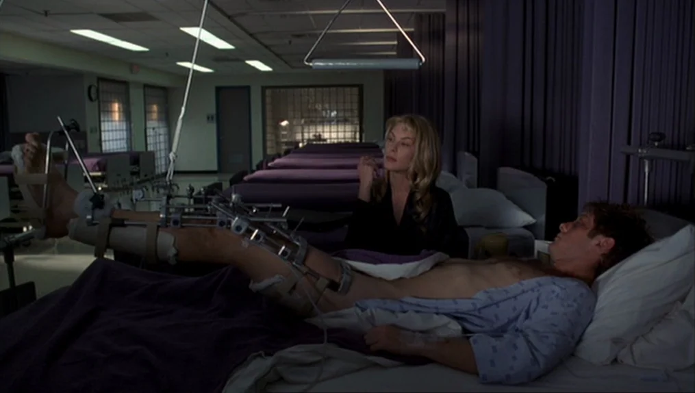 Crash (1996) movie still: medium — A dark hospital ward filled with multiple beds and purple privacy curtains. A man with a heavy metal leg brace…