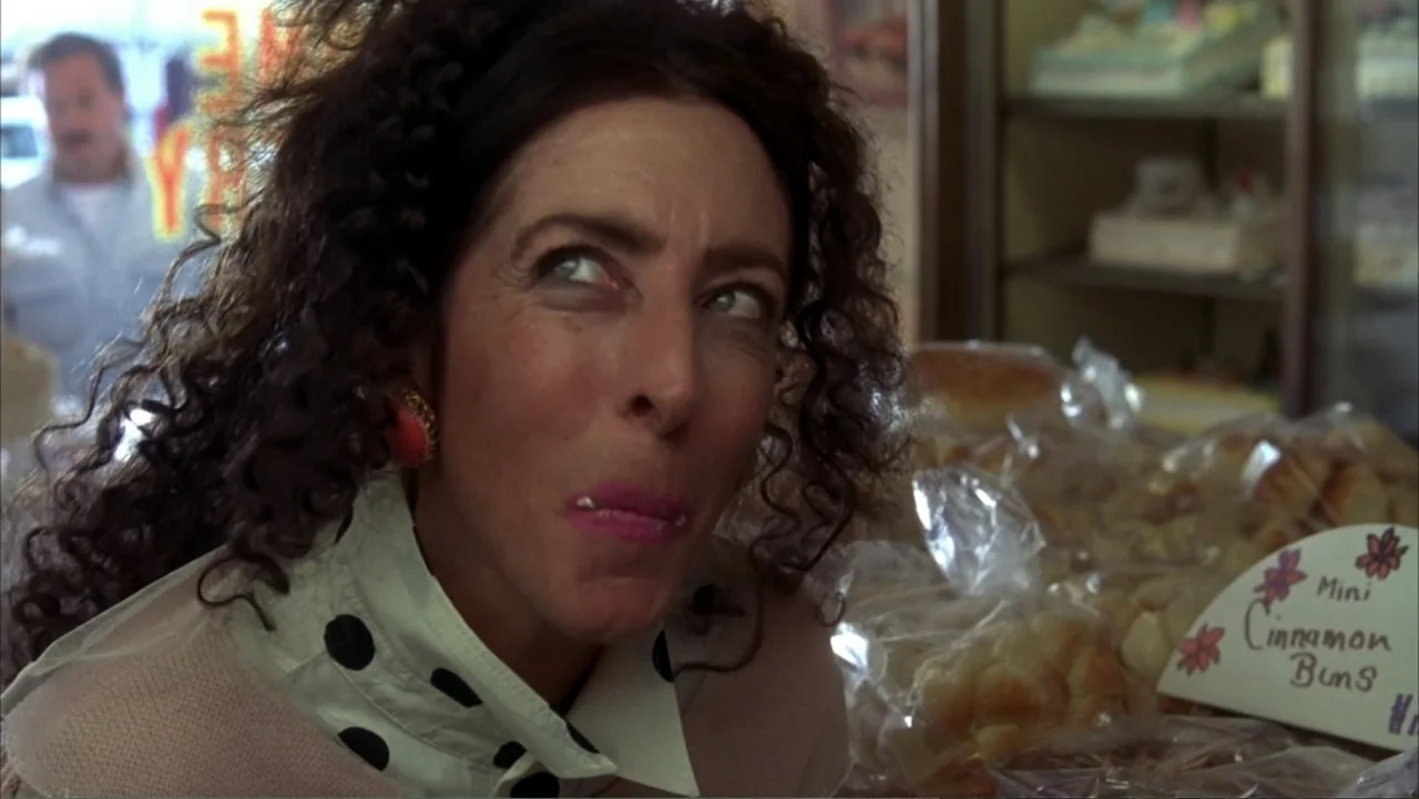 The Wedding Singer (1998) movie still: closeup — A bright bakery interior features a glass display case filled with baked goods. A woman with dark…