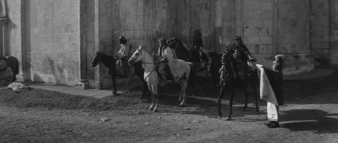 Andrei Rublev (1966) movie still: wide — A large stone masonry wall forms the backdrop of an outdoor dirt courtyard area. Multiple riders on…