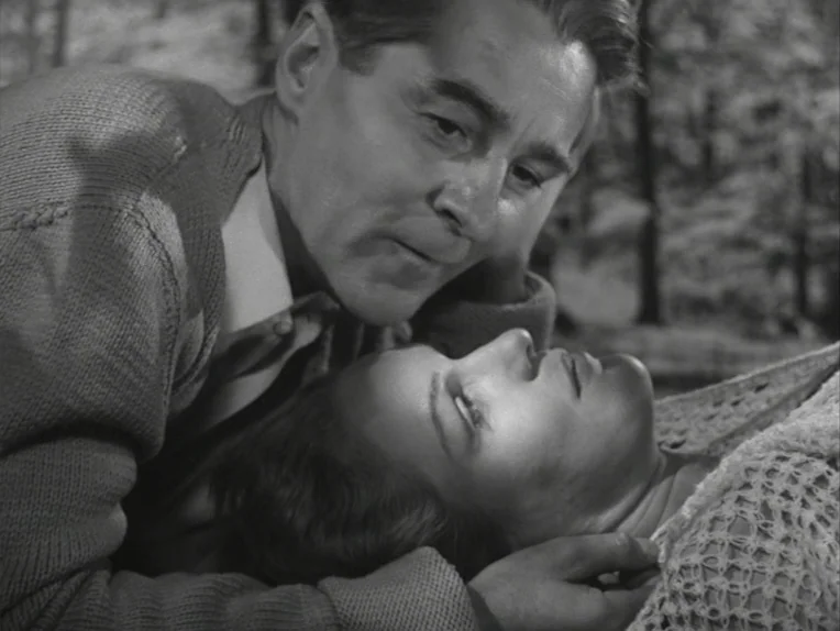 A Lesson In Love (1954) movie still: closeup — A man and woman are lying on the ground in a dense, treed area. The man leans over the woman while…