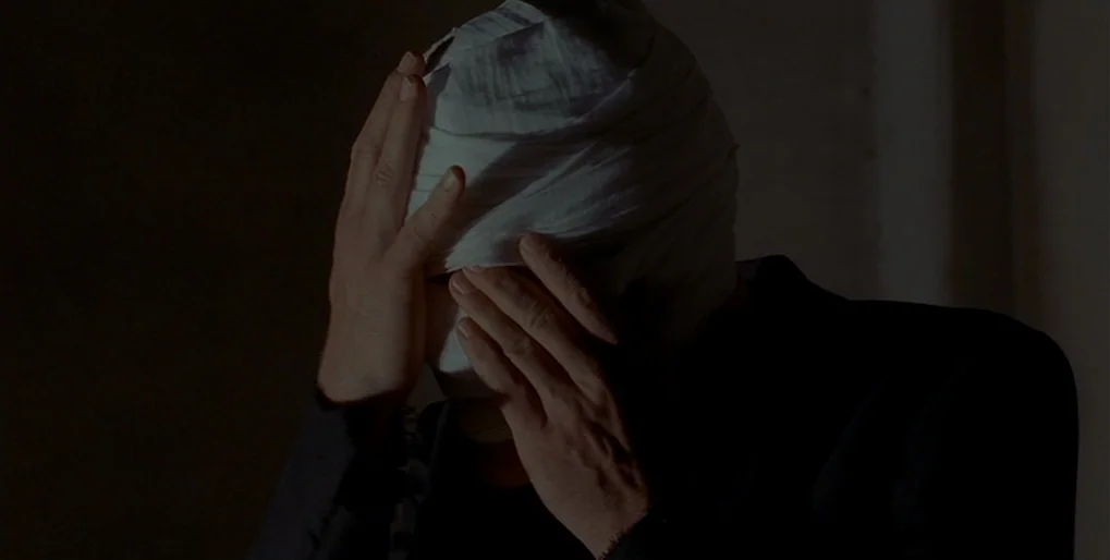 Dominion: Prequel to the Exorcist (2005) movie still: closeup — A dimly lit, dark setting frames a person with a bandaged head. The subject raises…