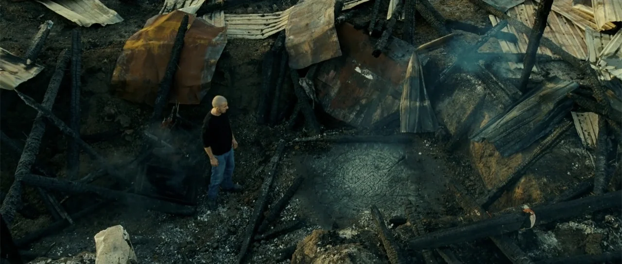 Blood Creek (2009) movie still: wide — The site of a burned structure features charred timber and metal sheets on dirt. A man in a black shirt…