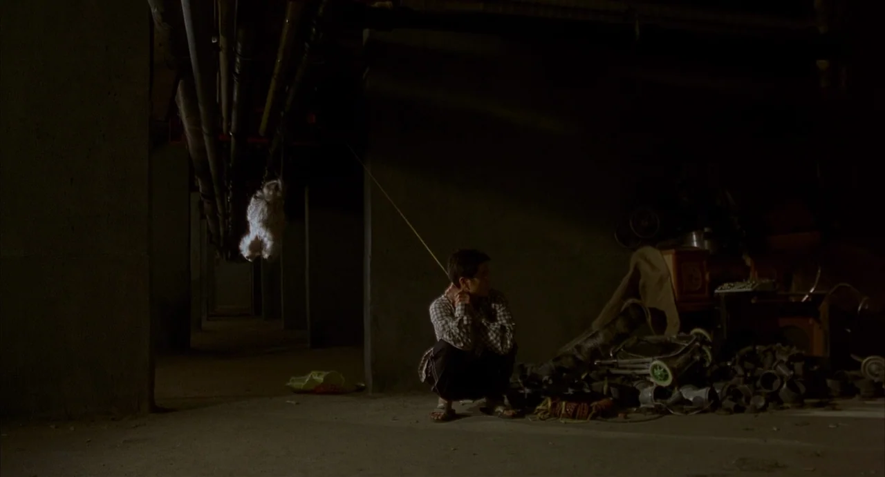 Barking Dogs Never Bite (2000) movie still: wide — A dark concrete basement filled with pipes and cluttered storage items. A man crouches on the…