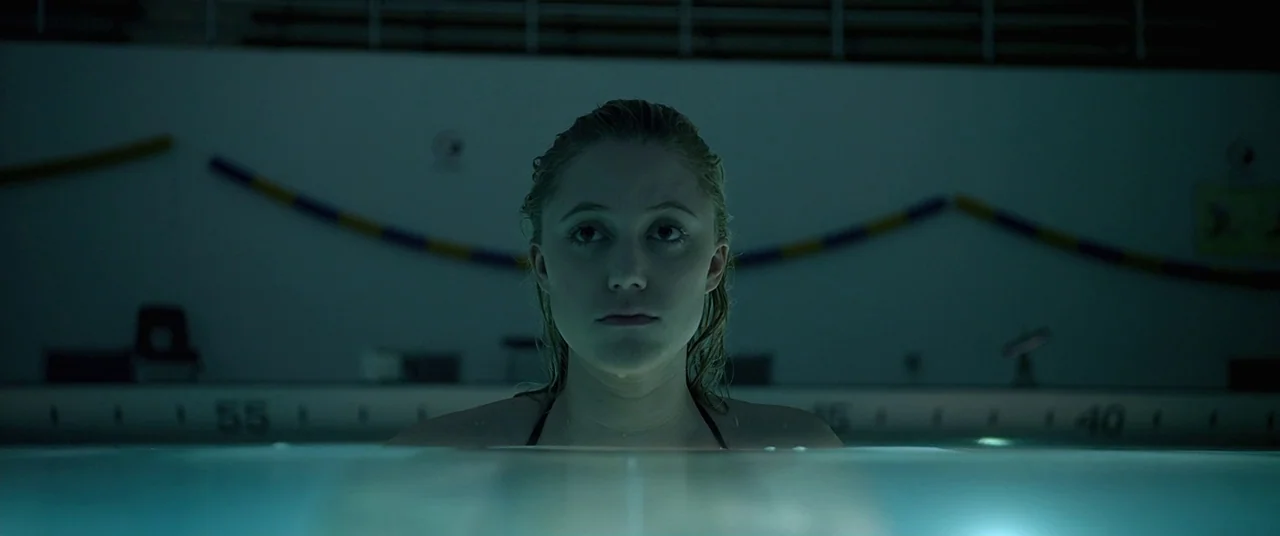 It Follows (2015) movie still: medium — A dimly lit indoor pool at night with murky blue water and tiled edges. A young woman with wet blonde hair…