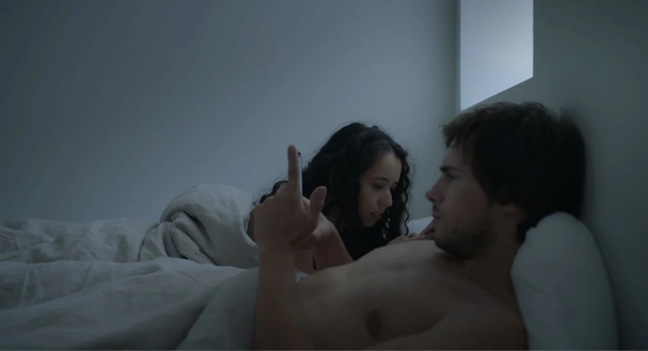 This Closeness (2024) movie still: medium — A dimly lit room at night features a couple resting in bed together. The man and woman look at the light…
