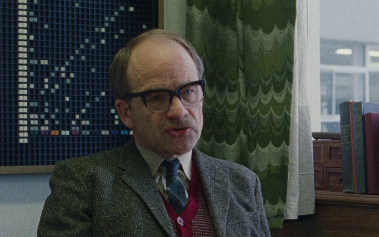 Education (2020) movie still: medium — An interior office space featuring a geometric wall display and green patterned curtains. A man in a blazer…