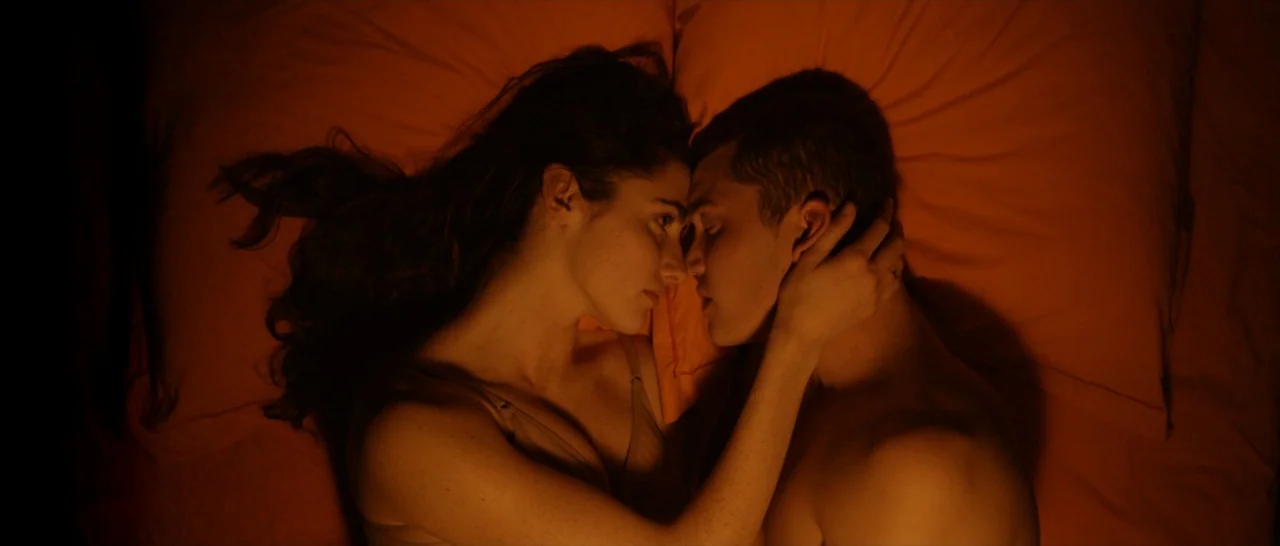 Love (2015) movie still: medium — A man and a woman lie face-to-face on vibrant orange bedding in a dim room. The woman reaches out with her hand to…