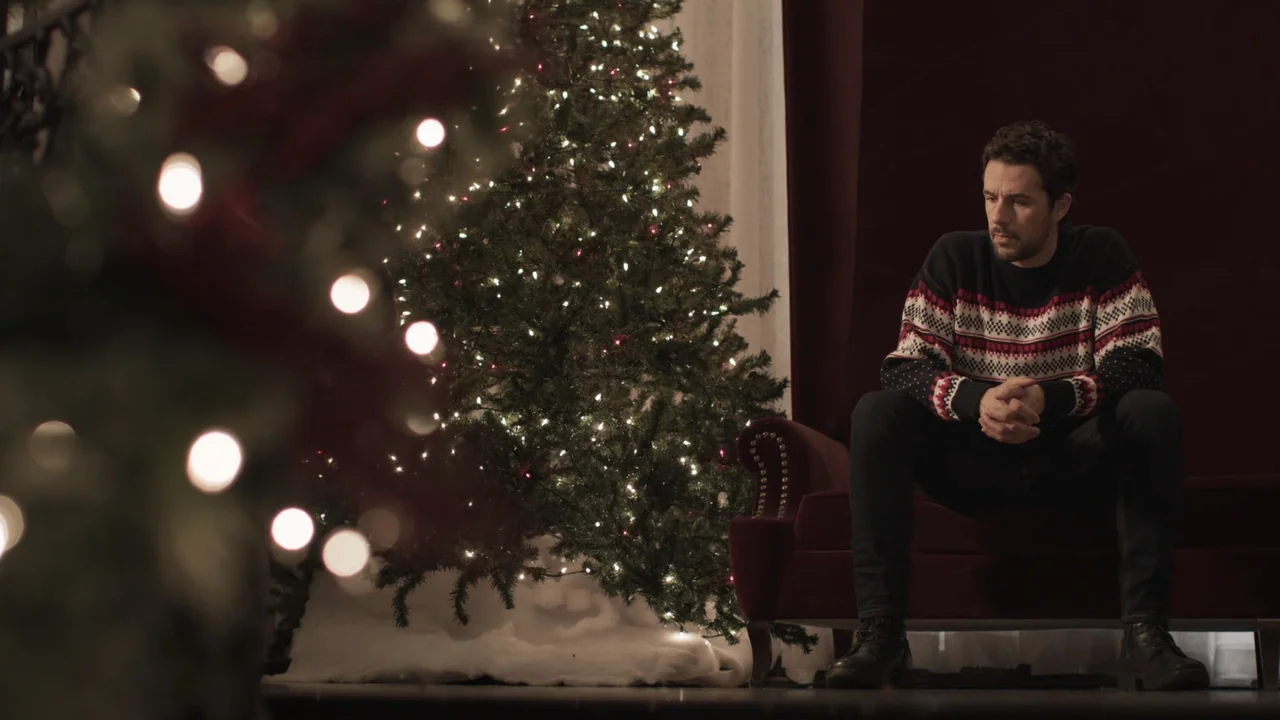 A Gingerbread Christmas (2022) movie still: medium — A man sits in an armchair next to a glowing, decorated holiday pine tree. Dark red curtains…