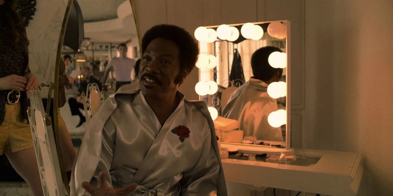 Dolemite Is My Name movie still: medium — A brightly lit vanity area features a mirror surrounded by numerous glowing round bulbs. The man in the…