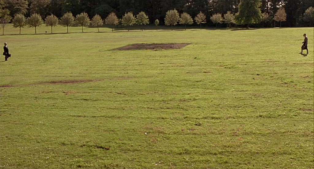 Trainspotting (1996) movie still: wide — A vast green public park features a line of evenly spaced trees in back. Two men walk across the open grass…