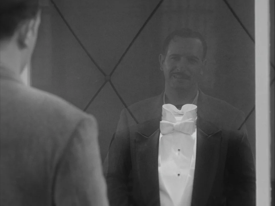 The Artist (2011) movie still: medium — A man adjusts his formal attire while looking at his reflection in a wall mirror. The mirror surface has a…