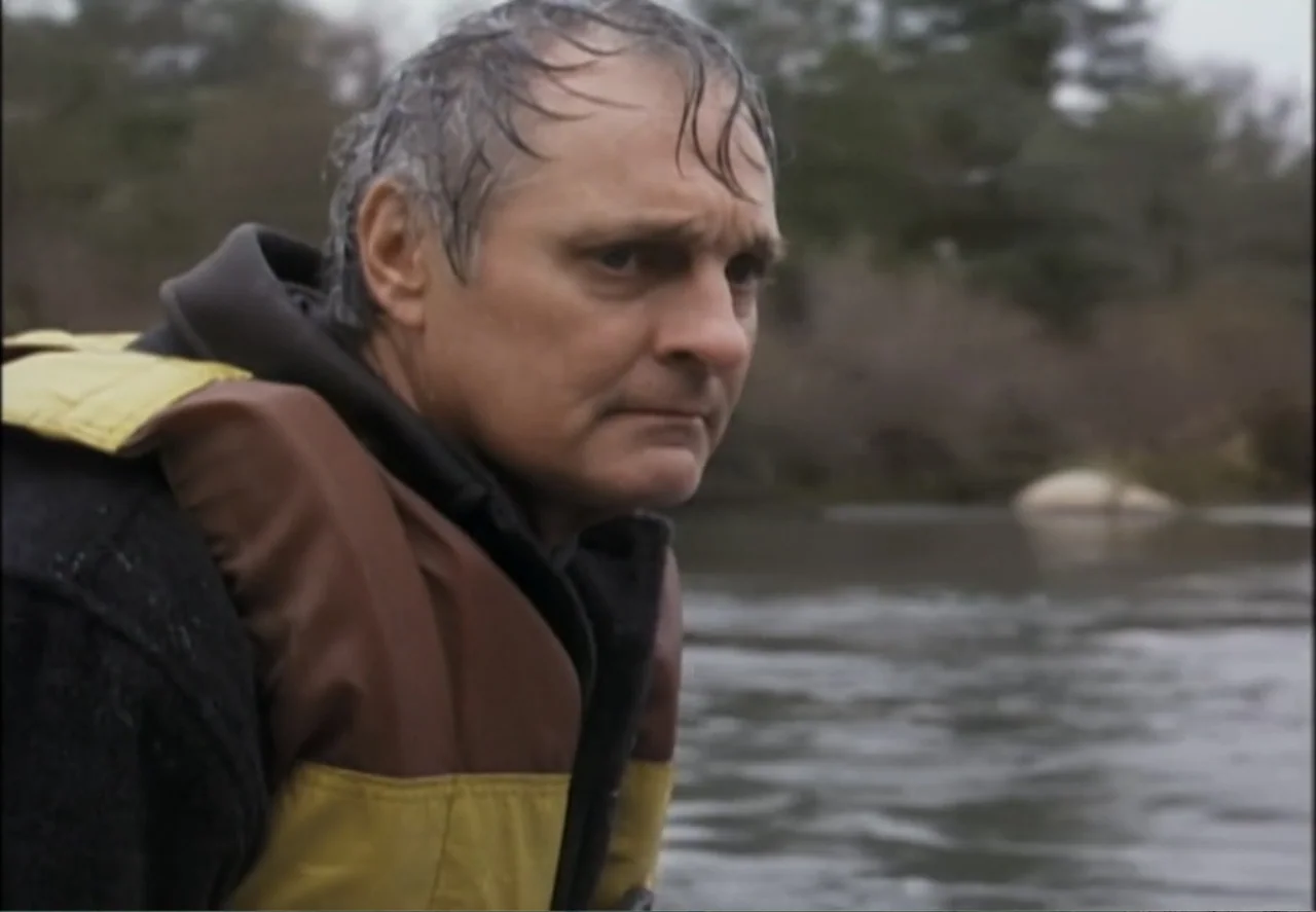 White Mile (1994) movie still: medium — A man stands near a gray, flowing river under an overcast sky. He is dressed in a life jacket and gazes…