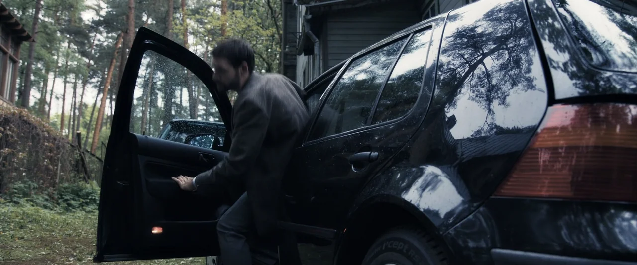The Good Neighbor (2022) movie still: medium — A dark sedan is parked on a wet grassy area near a wooden building. A man in a dark blazer steps out…