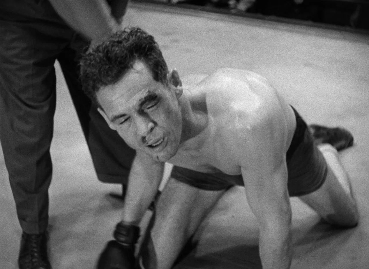 The Set-Up (1949) movie still: medium — A man kneels on the canvas surface within a brightly lit indoor arena. The boxer looks downward with visible…