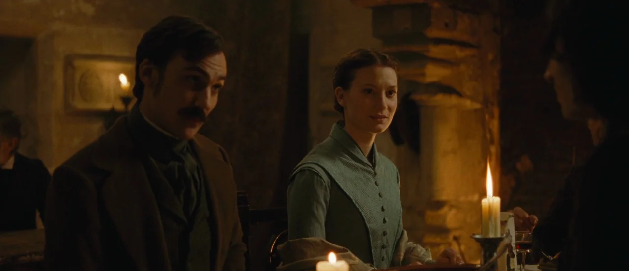 Madame Bovary movie still: medium — A dimly lit room with stone walls appears to be an 1800s dining area. Three people sit around a table…
