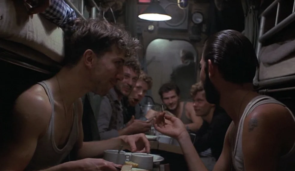 Das Boot (1981) movie still: medium — A cramped, metallic submarine bunk room filled with pipes and clustered bunks. Several young men sit around a…