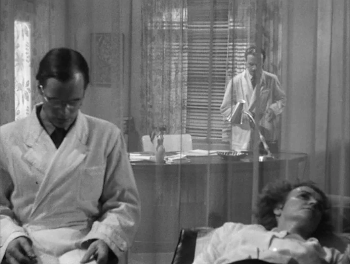Thirst (1949) movie still: medium — A sterile medical office features a desk, Venetian blinds, and sheer privacy curtains. A man in a lab coat sits…