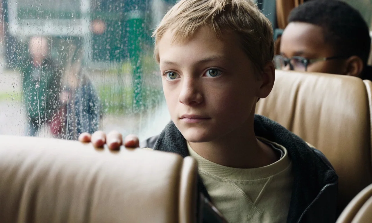 Close (2022) movie still: medium — A bus interior with tan leather headrests and a wet glass window. A young boy looks out the window while another…