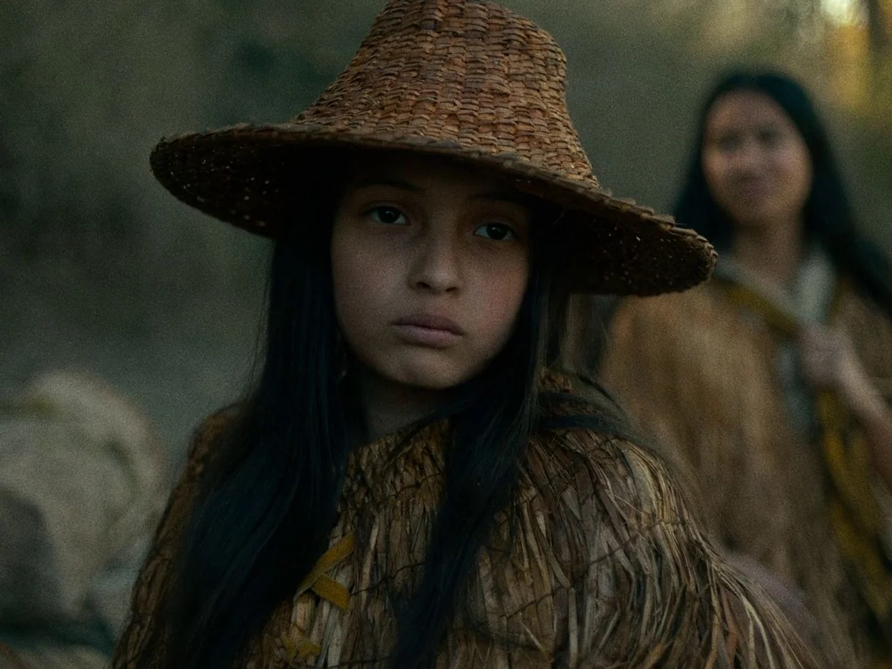 First Cow (2019) movie still: medium — A person in a woven straw hat stands outdoors in a dim wilderness setting. A second person is visible in the…