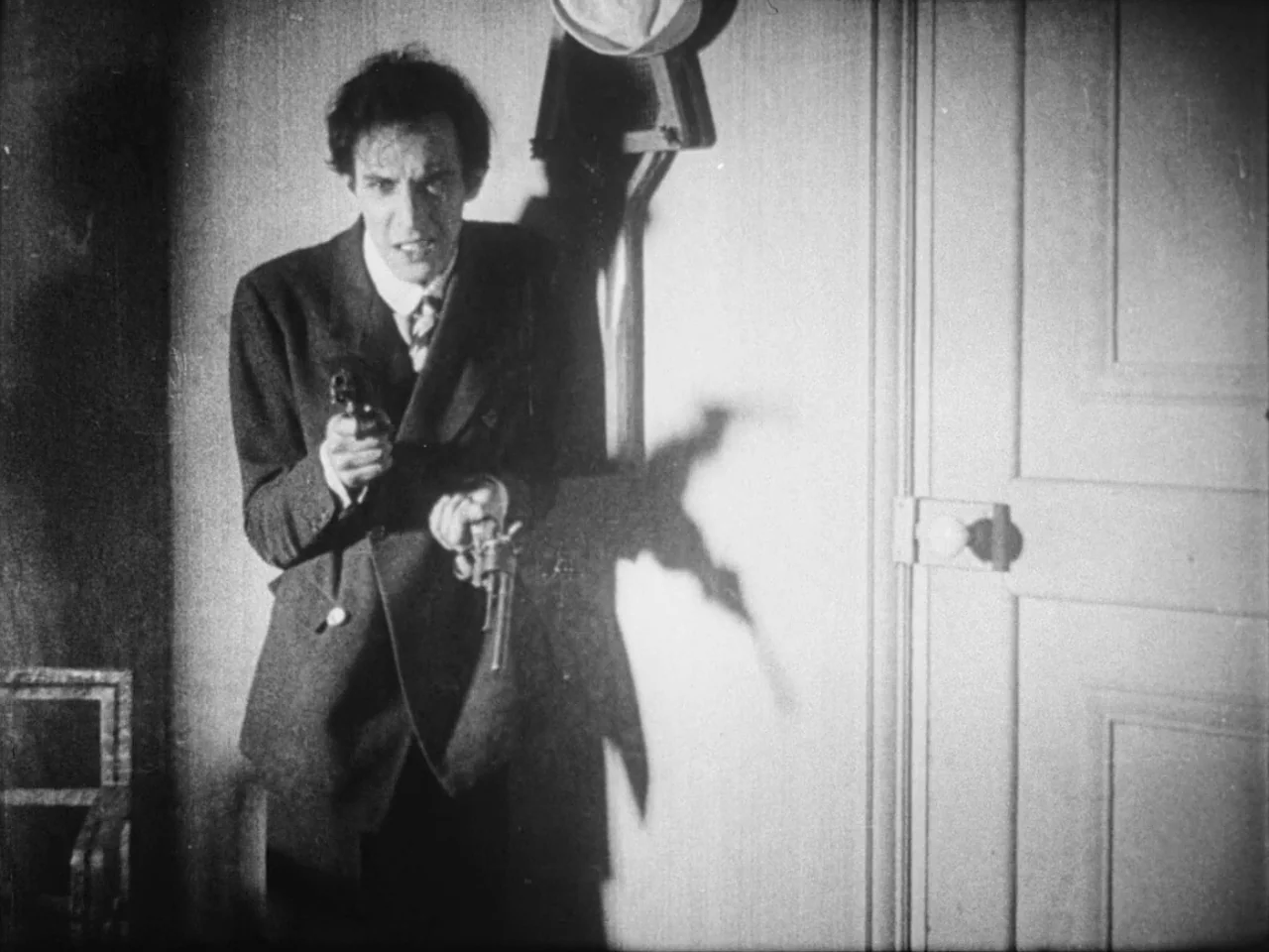 Un Chien Andalou (1929) movie still: medium — A man in a 1920s suit stands in a hallway beside a closed door. He holds two pistols leveled directly…