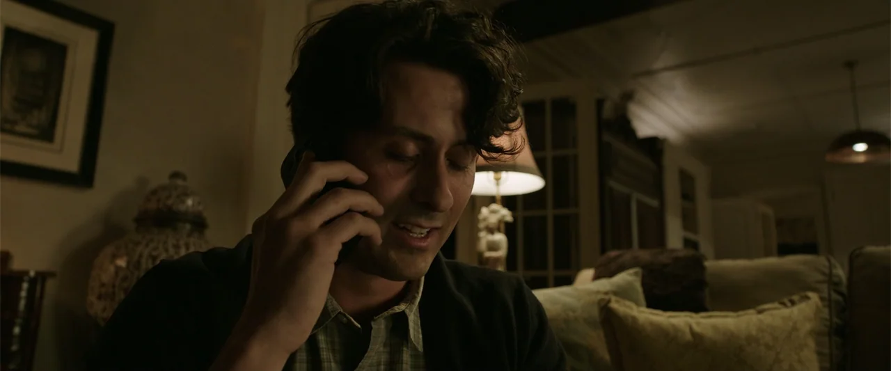 K.G.F: Chapter 2 (2022) movie still: closeup — A dimly lit room contains a sofa and a lit table lamp. A man sits while talking on his mobile phone.