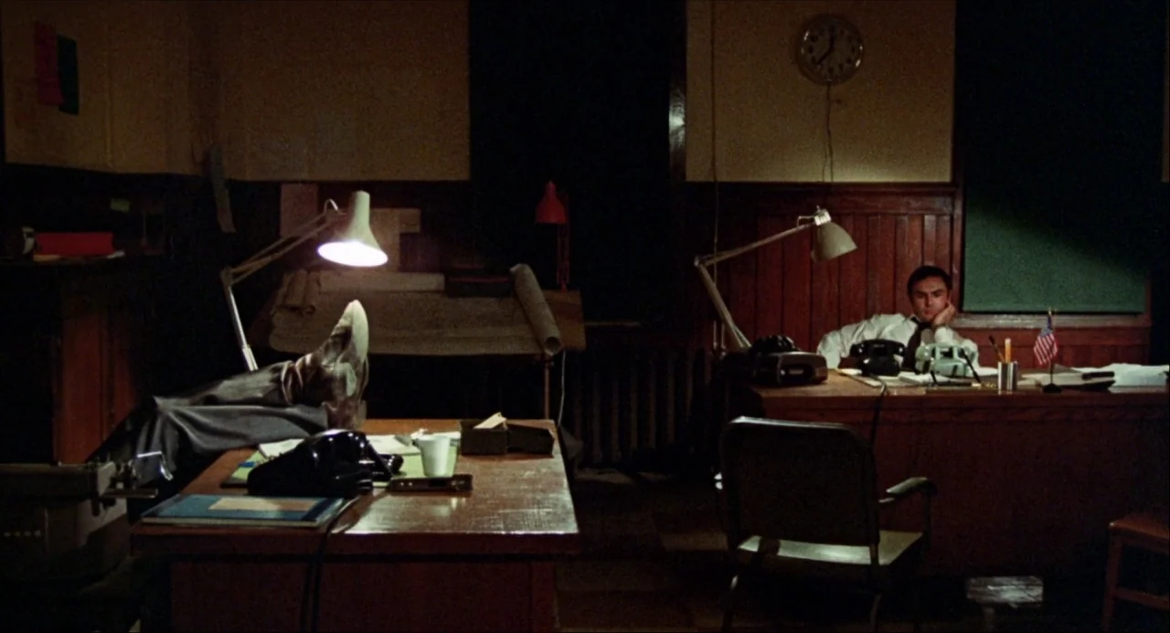 Black Christmas (1974) movie still: wide — A dimly lit office features dark wood paneling and two cluttered desks at night. A man sits at his desk…