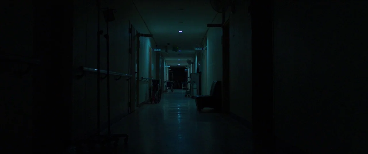 The Wailing (2016) movie still: establishing — A long, dimly lit hospital corridor stretches into the distance with doors on sides. Several medical…