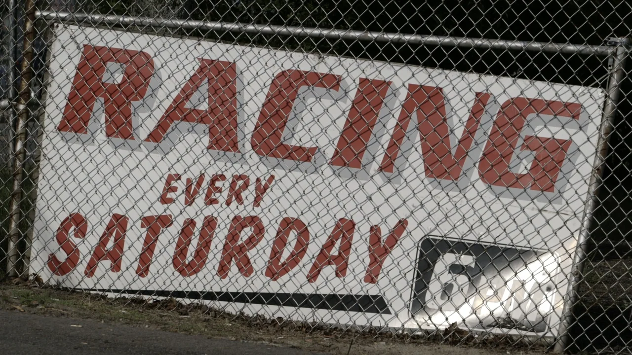 The Last Race (2022) movie still: medium — A white metal sign with red text advertising racing is attached to a fence. The sign reads racing every…